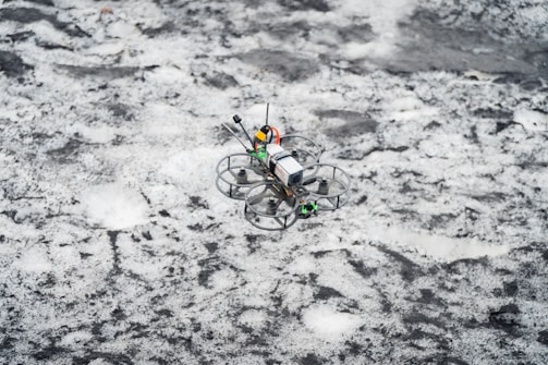 A small drone is flying over a snowy surface. The drone has a compact design with a camera and propellers protected by circular guards. The background consists of a snowy landscape with patches of dark ground and snow scattered unevenly.