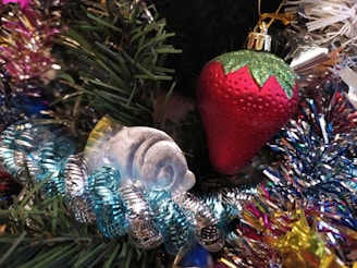 An assortment of festive decorations includes a red strawberry-shaped ornament with green glittered leaves, surrounded by glistening tinsel and artificial evergreen branches. A translucent fish ornament with blue iridescent detailing is nestled nearby, enhancing the holiday aesthetic.