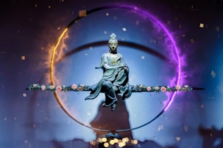 A statue of a serene figure, possibly a deity or Buddha, is suspended in the center of a vivid circular light ring. The ring displays a gradient of colors from fiery orange to mystical purple. The figure sits in a meditative posture on a horizontal rod adorned with delicate pink roses. Small confetti-like particles float around, contributing to an ethereal and spiritual atmosphere.