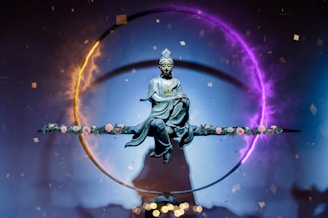 A statue of a serene figure, possibly a deity or Buddha, is suspended in the center of a vivid circular light ring. The ring displays a gradient of colors from fiery orange to mystical purple. The figure sits in a meditative posture on a horizontal rod adorned with delicate pink roses. Small confetti-like particles float around, contributing to an ethereal and spiritual atmosphere.