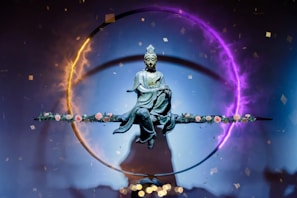 A statue of a serene figure, possibly a deity or Buddha, is suspended in the center of a vivid circular light ring. The ring displays a gradient of colors from fiery orange to mystical purple. The figure sits in a meditative posture on a horizontal rod adorned with delicate pink roses. Small confetti-like particles float around, contributing to an ethereal and spiritual atmosphere.