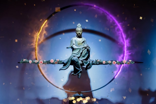 A statue of a serene figure, possibly a deity or Buddha, is suspended in the center of a vivid circular light ring. The ring displays a gradient of colors from fiery orange to mystical purple. The figure sits in a meditative posture on a horizontal rod adorned with delicate pink roses. Small confetti-like particles float around, contributing to an ethereal and spiritual atmosphere.