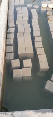 A series of rectangular cement blocks are partially submerged in a rectangular pool of water. The water is still, creating a reflective surface. Some round objects are floating on the water nearby, and there is a visible ledge surrounding the pool.