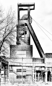 A black and white photograph of an industrial structure, likely a mining headframe, with prominent lines and geometric shapes. The structure is composed of metal and brick, featuring staircases and large windows. In the foreground, leafless tree branches create additional texture.