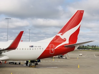 A passenger airplane on the tarmac with a distinctive red tail fin featuring a white kangaroo logo. The aircraft is parked next to an airport terminal, and there are several ground service vehicles nearby. The sky is overcast with soft, diffused lighting.