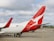 A passenger airplane on the tarmac with a distinctive red tail fin featuring a white kangaroo logo. The aircraft is parked next to an airport terminal, and there are several ground service vehicles nearby. The sky is overcast with soft, diffused lighting.