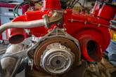 A detailed view of an exposed automotive engine with a bright red covering, showcasing various mechanical components such as exhaust pipes and a flywheel. The engine is situated in a workshop with tools and storage units in the background.