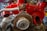 A detailed view of an exposed automotive engine with a bright red covering, showcasing various mechanical components such as exhaust pipes and a flywheel. The engine is situated in a workshop with tools and storage units in the background.
