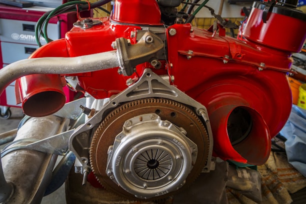 A detailed view of an exposed automotive engine with a bright red covering, showcasing various mechanical components such as exhaust pipes and a flywheel. The engine is situated in a workshop with tools and storage units in the background.