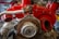 A detailed view of an exposed automotive engine with a bright red covering, showcasing various mechanical components such as exhaust pipes and a flywheel. The engine is situated in a workshop with tools and storage units in the background.