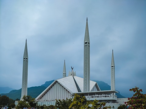 A modern, large white mosque with four tall minarets stands against a backdrop of misty, green mountains and a cloudy sky. The structure features angular, geometric designs with a prominent white roof and walls.