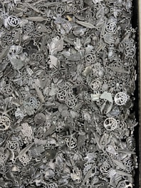 A dense assortment of small, metallic charms and trinkets that include various designs such as leaves, stars, hearts, and abstract shapes. The objects are closely packed together, showcasing intricate details and a variety of ornamental patterns.