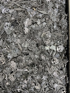 A dense assortment of small, metallic charms and trinkets that include various designs such as leaves, stars, hearts, and abstract shapes. The objects are closely packed together, showcasing intricate details and a variety of ornamental patterns.