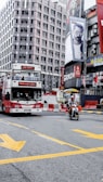 A busy urban street corner featuring a red double-decker bus with the words 'National Libra' on the front. A person rides a motorcycle in the foreground. The background includes tall buildings adorned with large advertisements and billboards. Bright yellow road markings are visible on the asphalt.