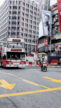 A busy urban street corner featuring a red double-decker bus with the words 'National Libra' on the front. A person rides a motorcycle in the foreground. The background includes tall buildings adorned with large advertisements and billboards. Bright yellow road markings are visible on the asphalt.