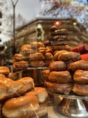 Stacks of glazed donuts are displayed on tiered metal trays in a shop window. The background shows reflections of buildings and trees, creating a cozy and inviting atmosphere.