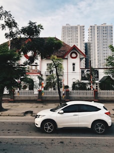 A white car is parked on a street in front of a historic building with red and white accents. Palm trees and a fenced garden are in front of the building. Tall modern skyscrapers rise in the background, contrasting with the traditional architecture. The sky is partly cloudy, and there are trees on the sidewalk.