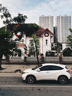 A white car is parked on a street in front of a historic building with red and white accents. Palm trees and a fenced garden are in front of the building. Tall modern skyscrapers rise in the background, contrasting with the traditional architecture. The sky is partly cloudy, and there are trees on the sidewalk.