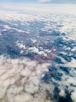 An aerial view of a landscape where patches of land are interspersed with bodies of water, creating a network of rivers or lakes. The scene is partially obscured by a layer of clouds that cover parts of the landscape. Urban areas and patches of greenery can be seen beneath the clouds, suggesting a mix of natural and developed areas.