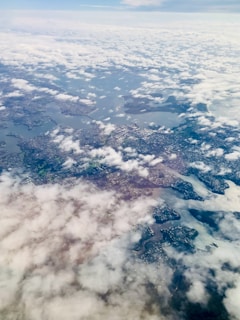 An aerial view of a landscape where patches of land are interspersed with bodies of water, creating a network of rivers or lakes. The scene is partially obscured by a layer of clouds that cover parts of the landscape. Urban areas and patches of greenery can be seen beneath the clouds, suggesting a mix of natural and developed areas.