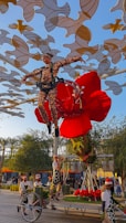 An acrobat wearing a patterned suit is suspended in the air by a harness, surrounded by a large red flower structure. There are multiple people on the ground wearing costumes, interacting with the floral installation. The background is filled with abstract golden and white shapes suspended overhead, set against a clear blue sky. Trees and a crowd are visible in the distance.