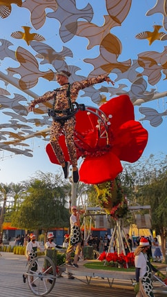 An acrobat wearing a patterned suit is suspended in the air by a harness, surrounded by a large red flower structure. There are multiple people on the ground wearing costumes, interacting with the floral installation. The background is filled with abstract golden and white shapes suspended overhead, set against a clear blue sky. Trees and a crowd are visible in the distance.