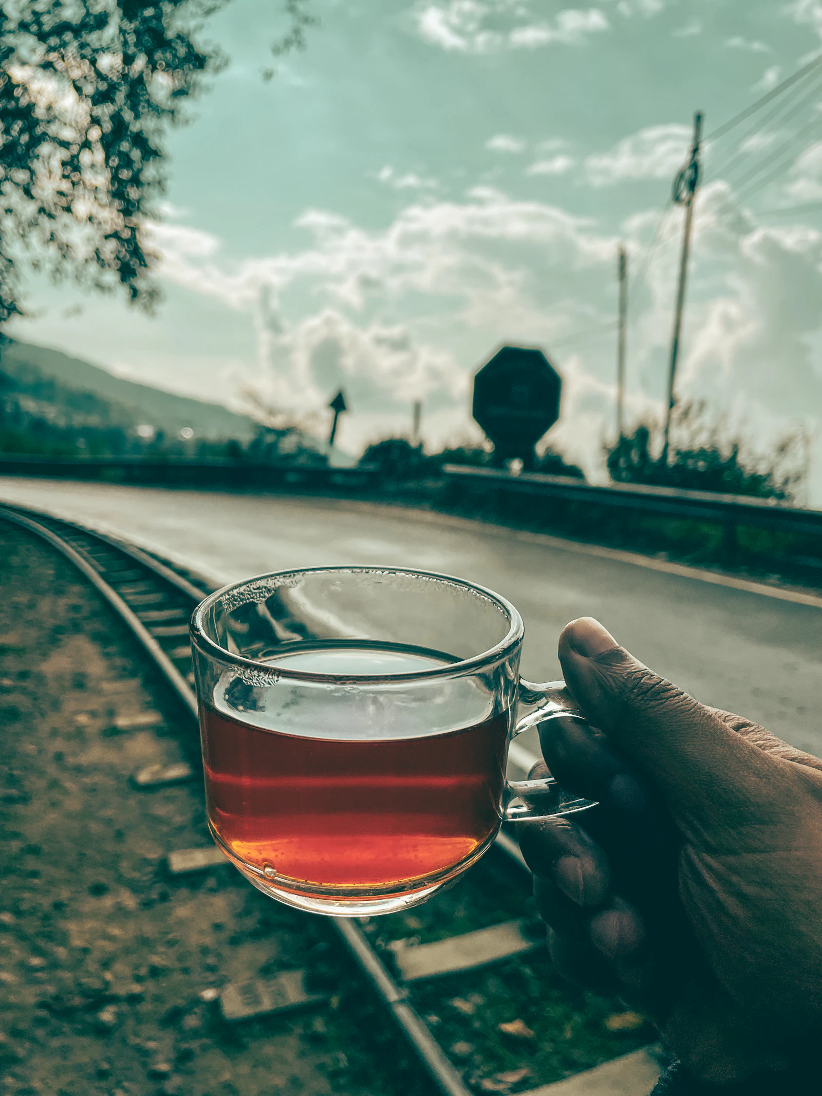 A glass of amber Darjeeling tea held up beside the toy train track and a quiet hill road under a pale sky