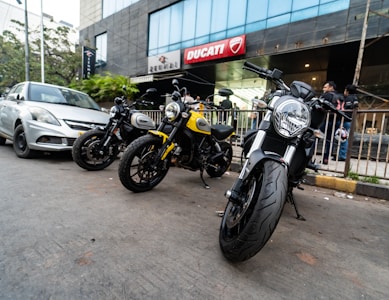 Several motorcycles are parked on a street near a modern building with a Ducati sign. A silver car is parked beside them, and a few people are visible in the background.