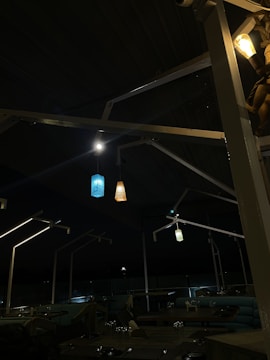 A dimly lit outdoor seating area with modern canopy structures and four hanging decorative lights. The lighting is subdued with a cozy ambiance, provided by blue, orange, and white pendants. Several tables with dining settings are visible in the foreground.