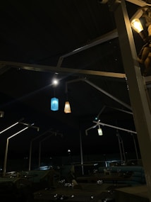 A dimly lit outdoor seating area with modern canopy structures and four hanging decorative lights. The lighting is subdued with a cozy ambiance, provided by blue, orange, and white pendants. Several tables with dining settings are visible in the foreground.