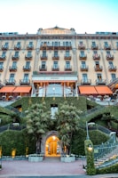 A grand hotel facade featuring multiple balconies and an elegant architecture. The building's entrance is surrounded by well-manicured greenery and palm trees. Stairs on both sides of the entrance have decorative metal railings and plants, creating a symmetrical look. The hotel name is displayed at the top of the building.