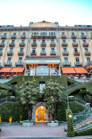 A grand hotel facade featuring multiple balconies and an elegant architecture. The building's entrance is surrounded by well-manicured greenery and palm trees. Stairs on both sides of the entrance have decorative metal railings and plants, creating a symmetrical look. The hotel name is displayed at the top of the building.