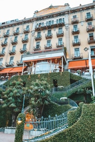 A grand hotel building with multiple rows of windows and balconies, featuring an ornate entrance surrounded by manicured greenery and palm trees. The facade has a classical design with decorative elements.