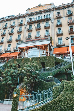 A grand hotel building with multiple rows of windows and balconies, featuring an ornate entrance surrounded by manicured greenery and palm trees. The facade has a classical design with decorative elements.