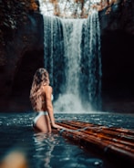 A person in swimwear stands on a bamboo raft in a river, facing a tall, cascading waterfall surrounded by lush vegetation.