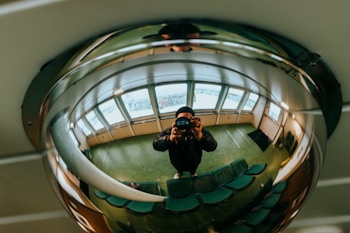 A person is taking a photo of themselves in a wide-angle view, captured in a reflective mirrored surface on the ceiling. The surroundings appear to be the interior of a room with green flooring, possibly a waiting area, featuring rows of green chairs and large windows with natural light streaming in.