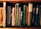 A collection of vintage books with worn spines, various titles, and richly textured covers standing upright on a wooden shelf. The books display a range of colors, including brown, blue, and green, showcasing intricate designs and aged paper.