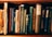 A collection of vintage books with worn spines, various titles, and richly textured covers standing upright on a wooden shelf. The books display a range of colors, including brown, blue, and green, showcasing intricate designs and aged paper.