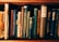 A collection of vintage books with worn spines, various titles, and richly textured covers standing upright on a wooden shelf. The books display a range of colors, including brown, blue, and green, showcasing intricate designs and aged paper.