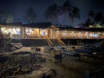 A nighttime beach restaurant featuring an open-air seating area with wooden tables and chairs. The structure is surrounded by palm trees and illuminated by warm, inviting lights. The sand is scattered with coconuts, adding to the tropical ambiance.