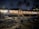 A nighttime beach restaurant featuring an open-air seating area with wooden tables and chairs. The structure is surrounded by palm trees and illuminated by warm, inviting lights. The sand is scattered with coconuts, adding to the tropical ambiance.