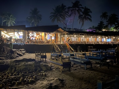 A nighttime beach restaurant featuring an open-air seating area with wooden tables and chairs. The structure is surrounded by palm trees and illuminated by warm, inviting lights. The sand is scattered with coconuts, adding to the tropical ambiance.