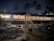 A nighttime beach restaurant featuring an open-air seating area with wooden tables and chairs. The structure is surrounded by palm trees and illuminated by warm, inviting lights. The sand is scattered with coconuts, adding to the tropical ambiance.