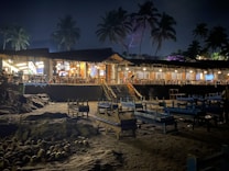 A nighttime beach restaurant featuring an open-air seating area with wooden tables and chairs. The structure is surrounded by palm trees and illuminated by warm, inviting lights. The sand is scattered with coconuts, adding to the tropical ambiance.