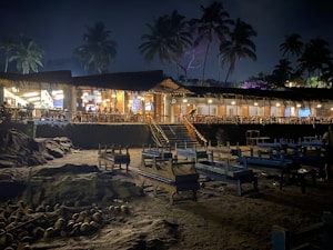 A nighttime beach restaurant featuring an open-air seating area with wooden tables and chairs. The structure is surrounded by palm trees and illuminated by warm, inviting lights. The sand is scattered with coconuts, adding to the tropical ambiance.