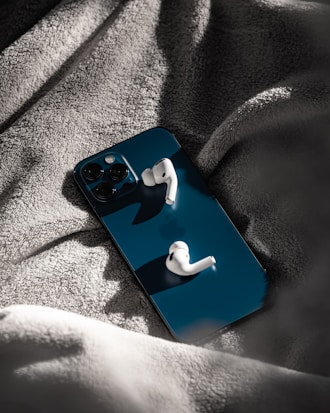 A smartphone with a reflective dark blue back surface is placed on a soft, gray fabric. Resting on top of the phone are wireless earbuds, casting distinct shadows.