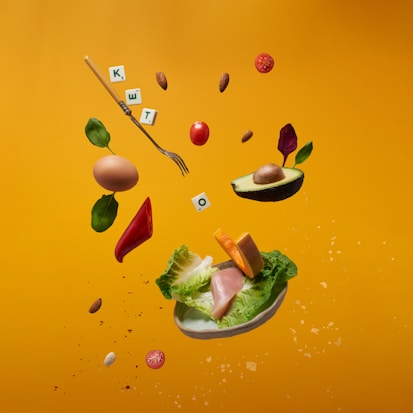 A vibrant display of floating food items includes an avocado half, a brown egg, cherry tomatoes, a slice of red bell pepper, almonds, and leafy greens. Among them, a fork and letter tiles spelling 'KETO' are interspersed. The background features a warm, mustard yellow color.