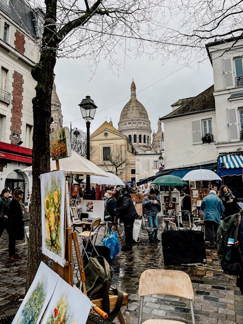 A bustling outdoor art market in an urban setting, with paintings displayed on easels and buildings surrounding the square. People mill about under umbrellas on a wet cobblestone street, with a prominent cathedral dome in the background.