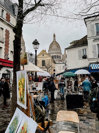 A bustling outdoor art market in an urban setting, with paintings displayed on easels and buildings surrounding the square. People mill about under umbrellas on a wet cobblestone street, with a prominent cathedral dome in the background.