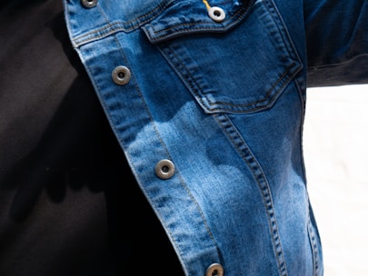 A close-up view of a person wearing a denim jacket with metal buttons and a black shirt underneath. The denim fabric shows various shades of blue, with visible stitching and a pocket on the chest. There are shadows cast on the jacket, indicating natural light.